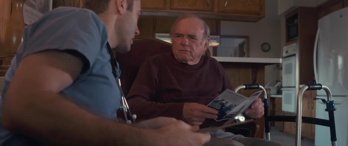 A male nurse in a blue shirt shows a brochure to an elderly man sitting in a chair with a walker nearby in a cozy woodpaneled room