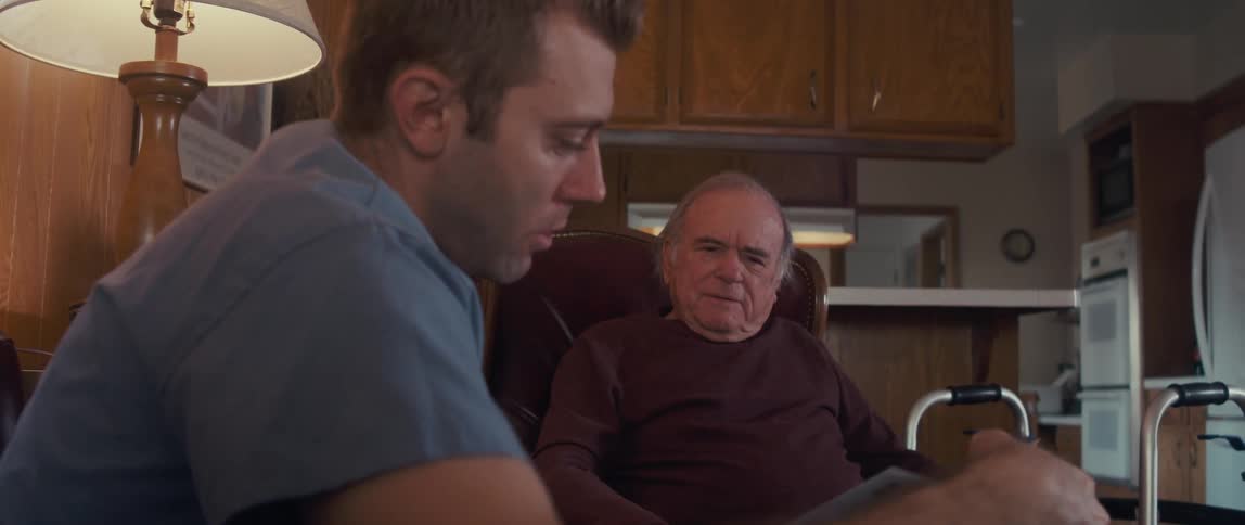 A male nurse in a blue shirt shows a brochure to an elderly man sitting in a chair with a walker nearby in a cozy woodpaneled room