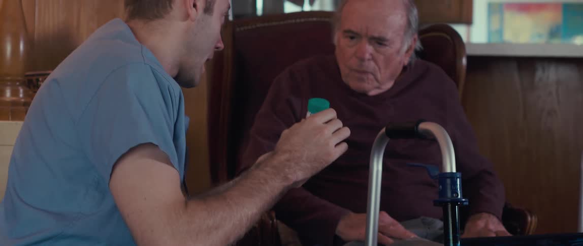 A male nurse in a blue shirt shows prescription pill bottles to an elderly man sitting in a chair with a walker nearby in a cozy woodpaneled room