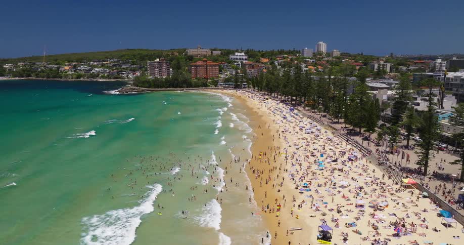A bustling beach on a sunny day where crowds of people are enjoying various activities like sunbathing swimming and strolling along the shore while clear blue water with gentle waves laps against the sandy beach