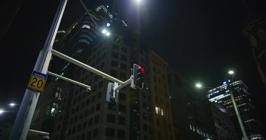 A nighttime street scene with a traffic light glowing red in the foreground and towering skyscrapers illuminated in the background