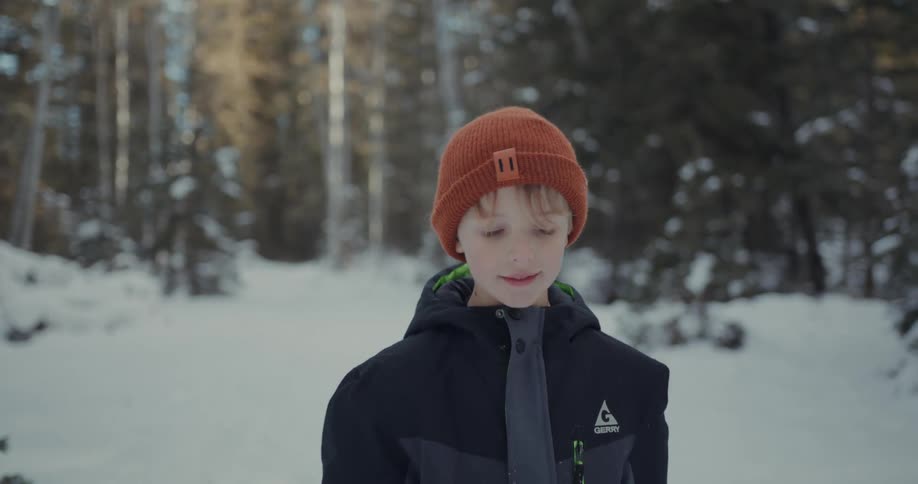 A boy wearing an orange beanie and a dark jacket walking on a snowy path near a forest