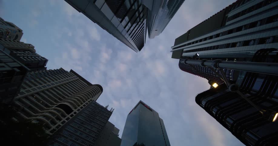 A cluster of tall buildings with varying architectural styles silhouetted against a cloudy early evening sky with a street lamp illuminated in the foreground