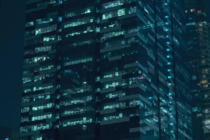 A skyscraper at night with the facade illuminated by a grid of flickering lights from the windows creating a dynamic pattern against the dark night sky