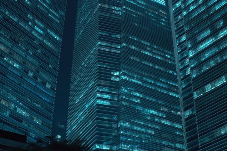 A cluster of towering skyscrapers bathed in a cool blue light that flickers rhythmically casts a mesmerizing glow against the dark night sky as their glass facades reflect the citys illumination