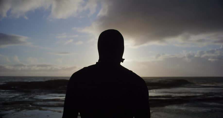 A silhouette of a person stands on a rocky shoreline facing the ocean with a dramatic sky featuring clouds and a bright sun