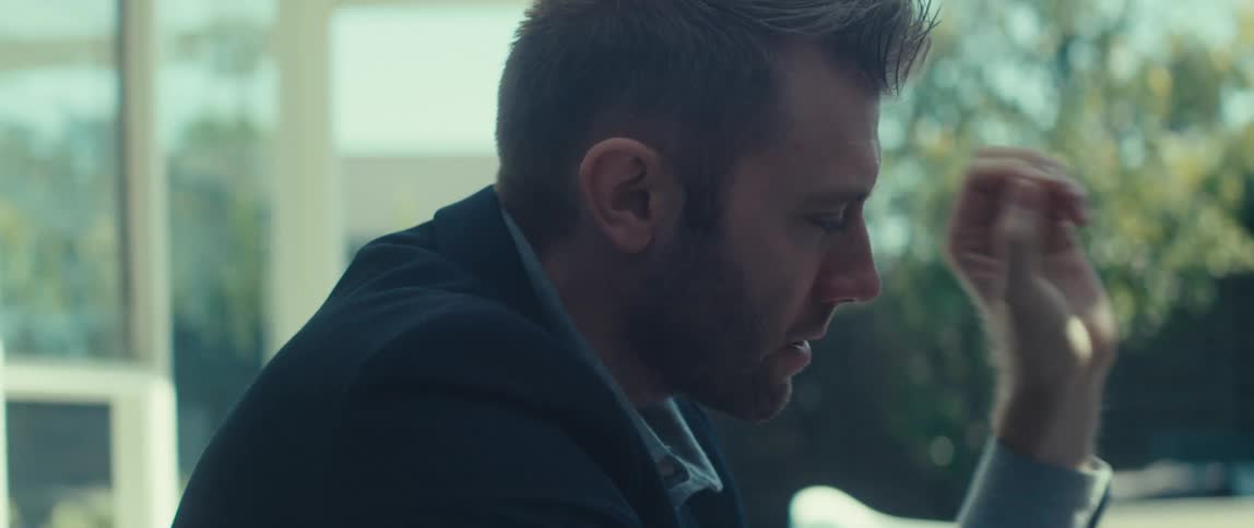 A person in a dark suit jacket and lightcolored shirt sits in the back yard talking