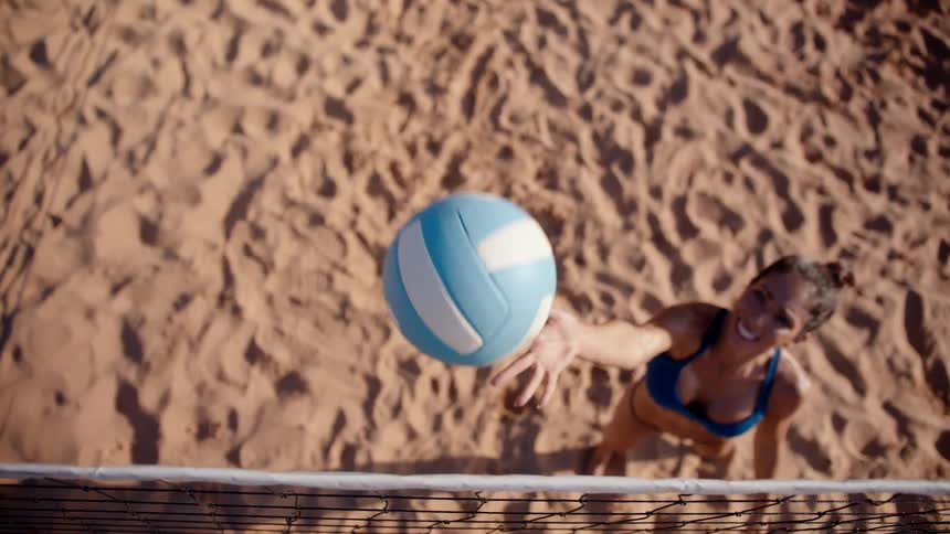 A smiling woman in a bikini is throwing a volleyball up in the air and she is on a sand volleyball court