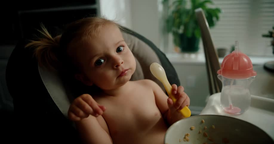 A young child is sitting in a high chair holding and nibbling on a spoon