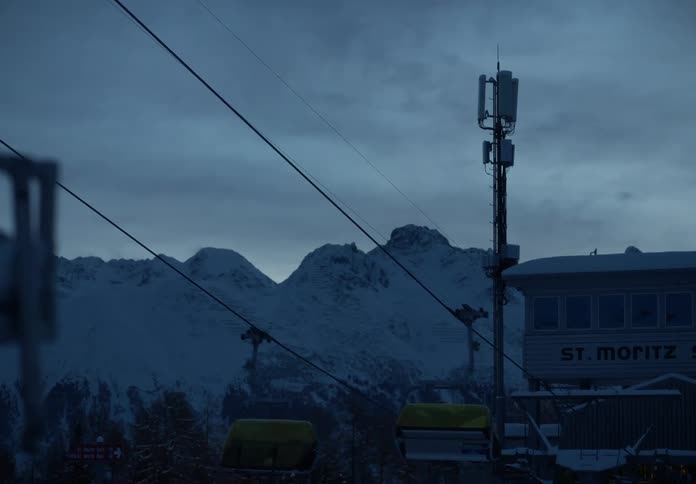 A ski lift chair gliding through a twilight sky with a snow covered mountain range in the background and a cell tower standing tall