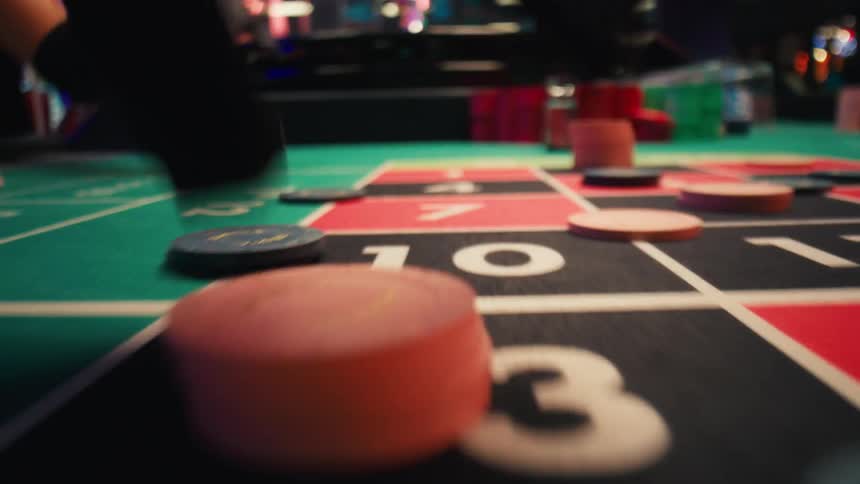 A hand placing a bet on a roulette table in a casino with other hands visible placing chips around the board