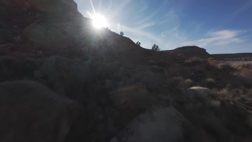 A rocky landscape with the sun shining brightly over a hill casting light rays across a clear blue sky with clouds
