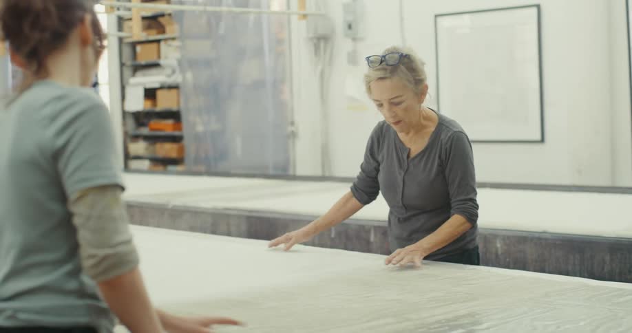 An older woman wearing glasses working on a project involving a large sheet of plastic or fabric while another person stands nearby observing in a workshop with natural light filtering through large windows