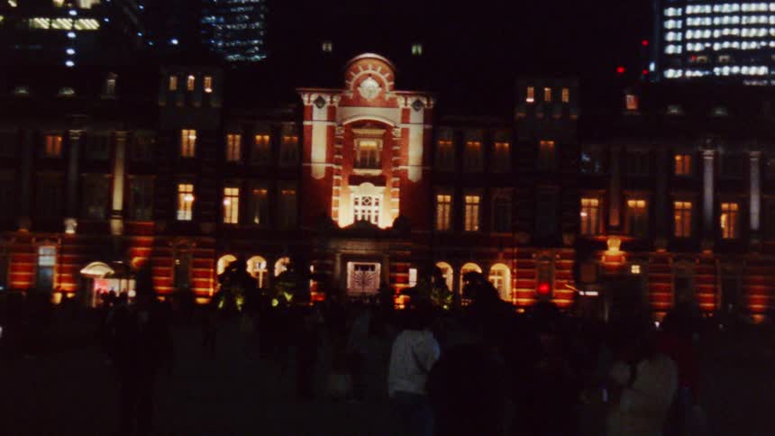 The ornate facade of a historic building glows with light casting a warm glow over a crowd of people gathered in the square below