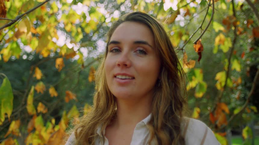 A woman with long hair is standing outdoors in front of a background of trees with yellow and green leaves