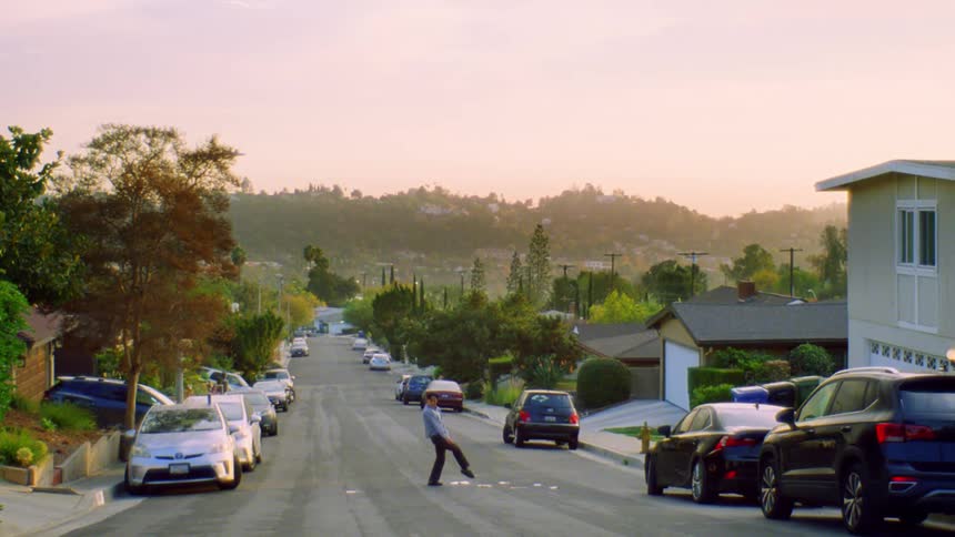 A person is dancing on a residential street lined with parked cars on both sides under a pinkish sky