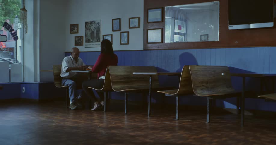 A man and a woman sit at a table in a small bluewalled diner with wooden chairs and framed pictures on the wall