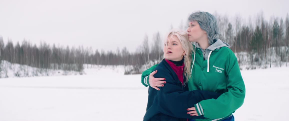 Two young women embrace as they walk in a snowy area with trees surrounding them