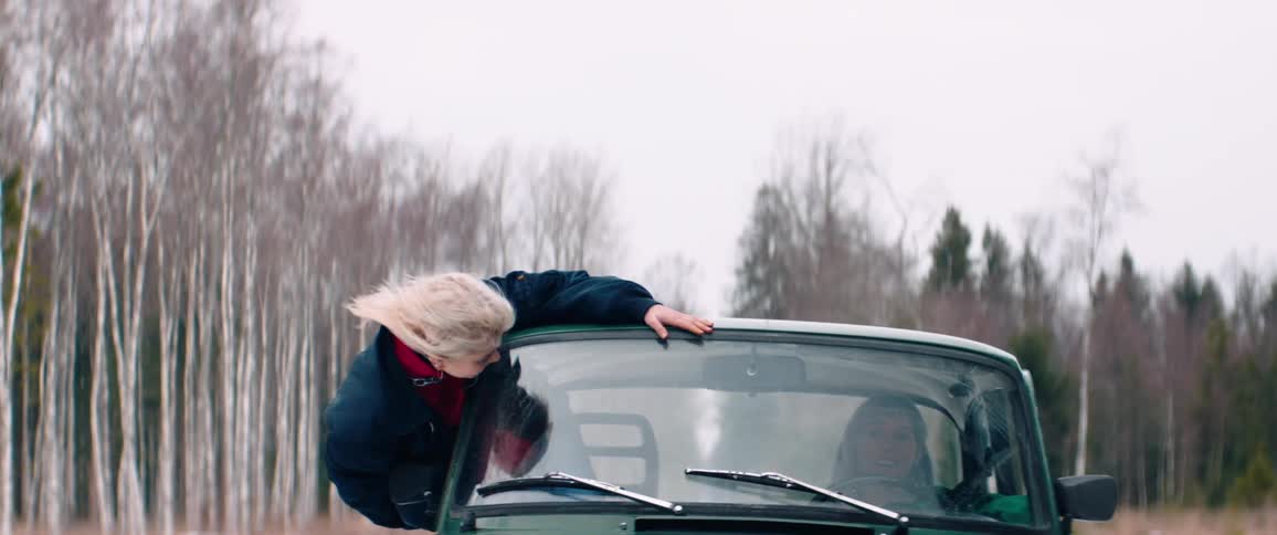 A woman is leaning out of the drivers side window of a green car driving through a snowy forested area