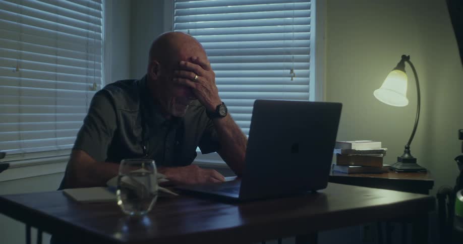 Elderly man sitting at an office desk in front of a laptop and crying