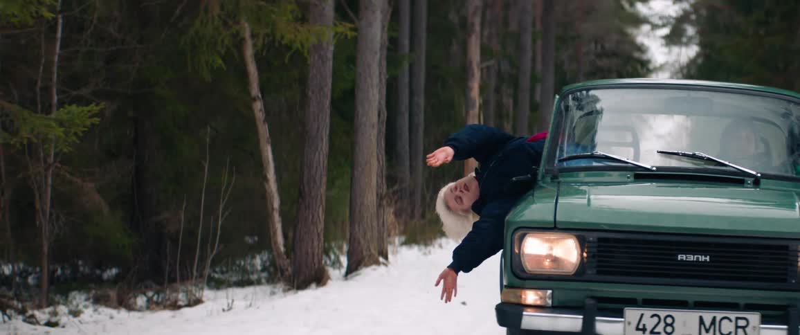 Classic vehicle moving through a snowy area surrounded by forests and a woman enjoying it with her body out of the window