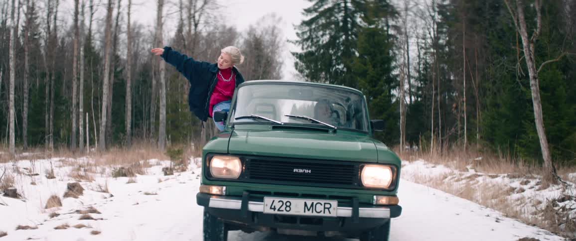 Green classic car traveling on a snowy road and a woman with her body out enjoying the vibe