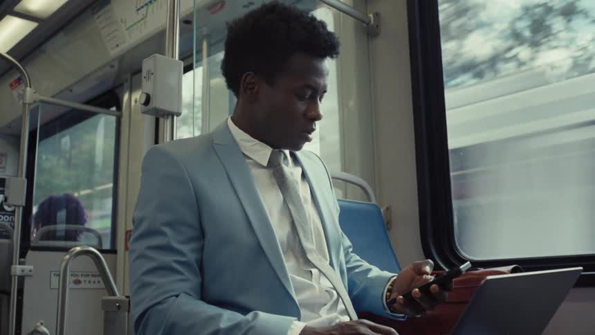A man in a suit answers a smartphone call while using a laptop on a train
