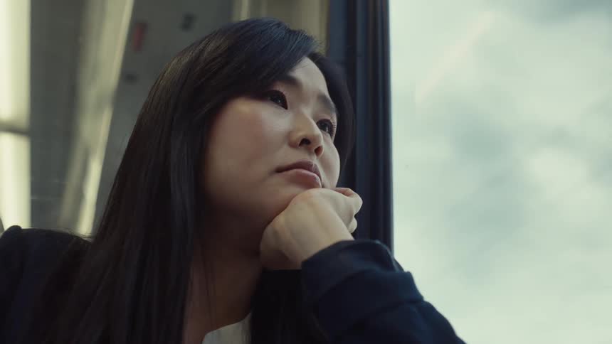 A woman with long dark hair is sitting by a window on a train resting their chin on their hand with a view of the sky