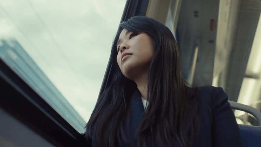 A woman with long black hair resting her head against a train window looking pensive as the cityscape reflects outside