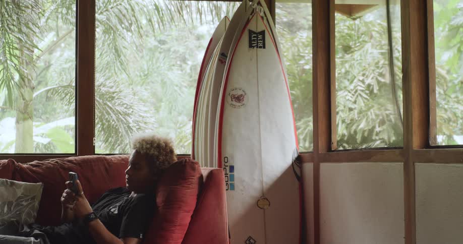 Man reclining on a couch near surfboards uses his smartphone