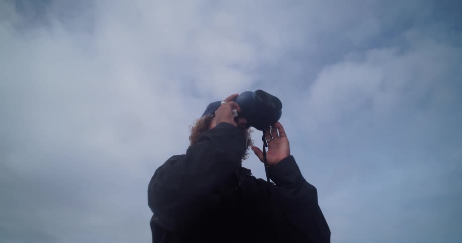An adult is standing outdoors while looking through binoculars on a cloudy day