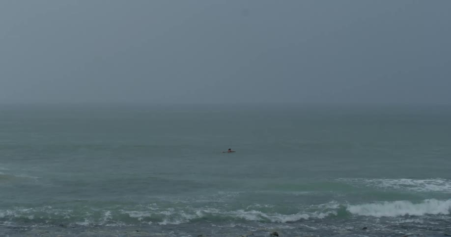 A lone surfer floats in the vast grey choppy ocean under a misty sky