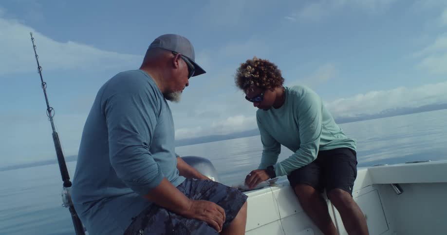 A man instructs a younger man about how to string a fish on a boat