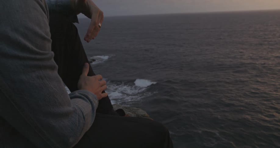A man taps on the side of his leg while sitting on a cliff by the ocean
