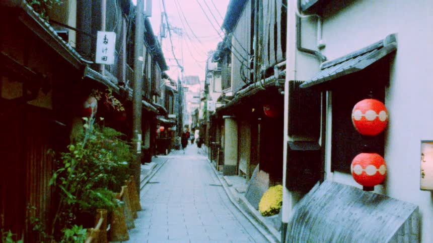 A narrow traditional japanese street lined with wooden buildings red lanterns and potted plants with overhead power lines and a few people walking in the distance