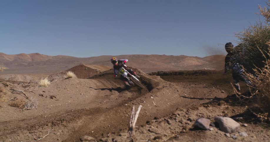 Two motorcyclists race dirt bikes down separate dirt tracks during the day