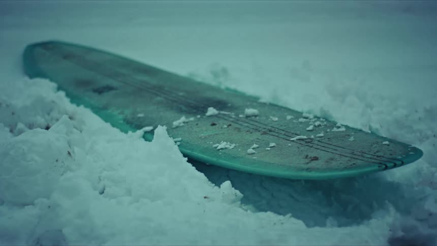 A surfboard lies on a snow covered surface with a cool bluish tint to the scene