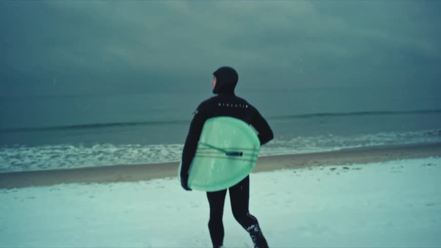 A man in a wetsuit carries a light blue surfboard through the snow on the beach near the sea under a cloudy sky