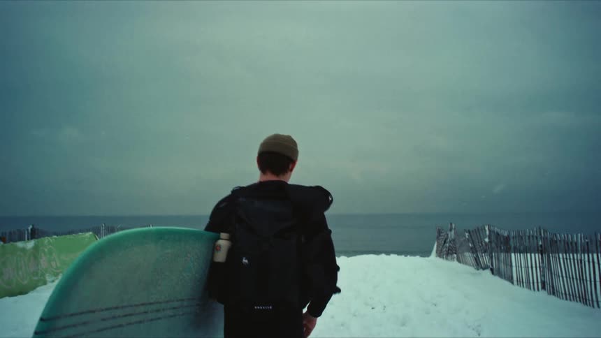 A person is walking towards the ocean on a snowcovered path carrying a surfboard under one arm with a wooden fence on the right