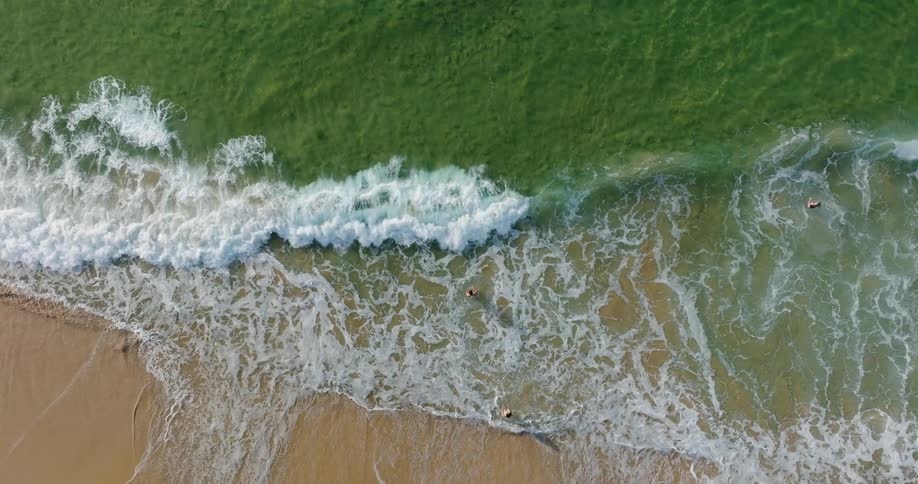 An aerial view shows waves crashing onto a sandy beach with people swimming in the water near the shore