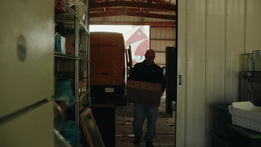 A man is carrying a box inside a barn with a delivery van parked at the entrance and a red barn is visible in the background