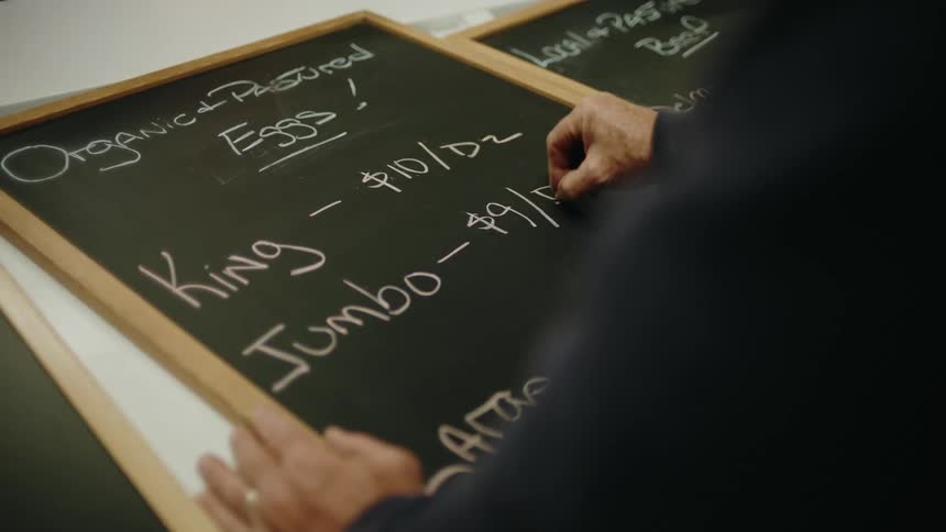 A person is writing on a chalkboard that lists prices for organic pastured eggs