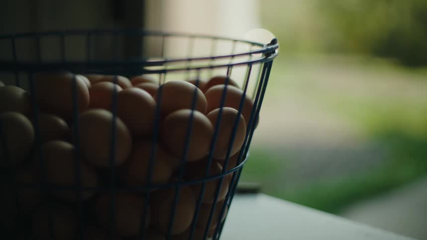 A wire basket filled with brown eggs placed on a white surface with a blurred outdoor background visible through a window
