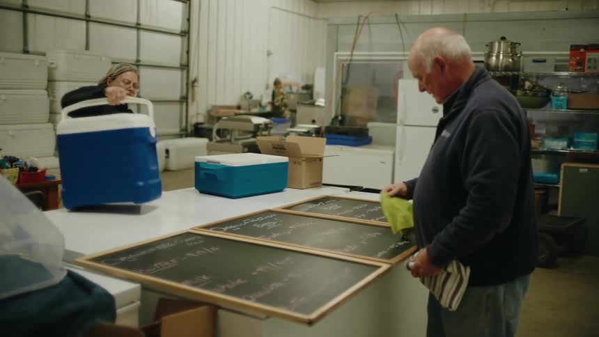 A woman carries in coolers as a man cleans a chalk board