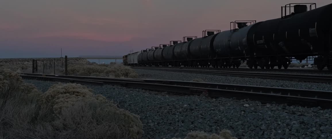A long train of black tanker cars on a railway track during sunset in a barren rocky rural landscape