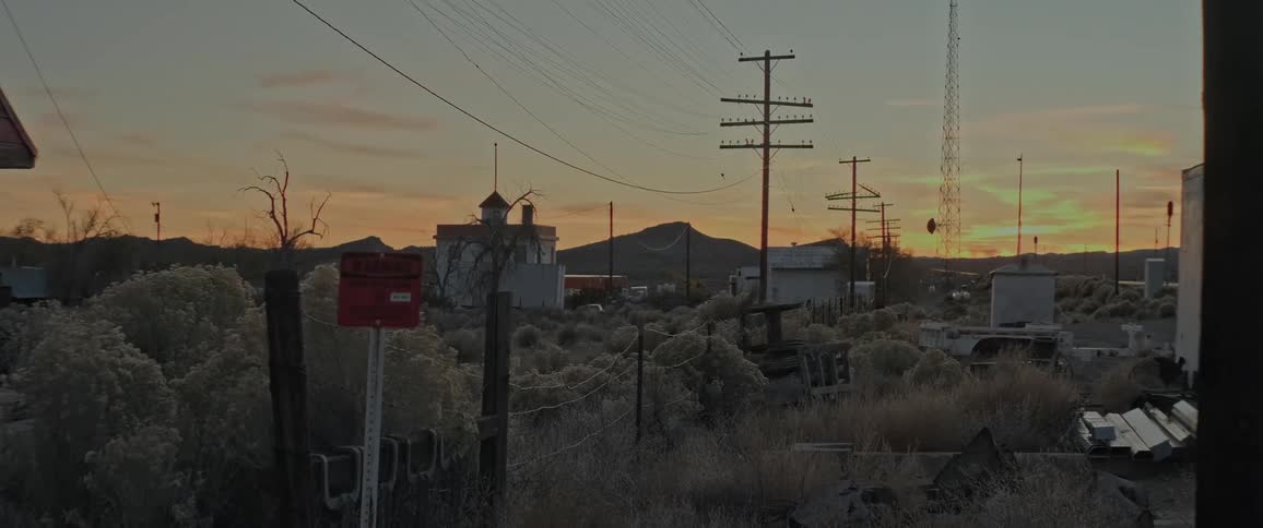 The setting sun casts a warm glow over a desolate landscape illuminating a cluster of abandoned buildings and power lines