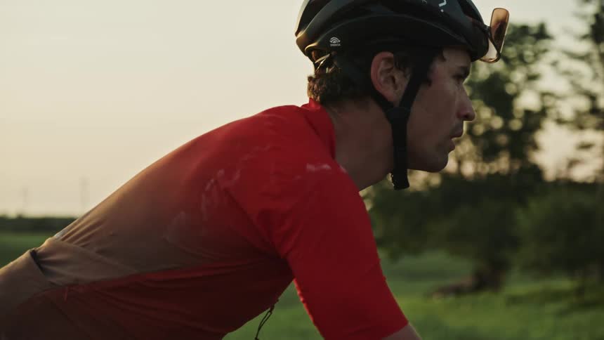Follow of a male road cyclist at sunset riding along a winding gravel road surrounded by lush green vegetation and rolling hills and in a rural landscape