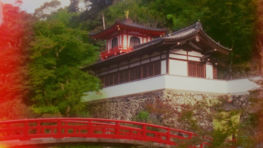 A traditional japanese temple with a red roof and white walls sits perched on a hillside overlooking a vibrant red bridge