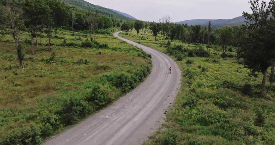 Drone shot of a lone cyclist riding along a winding gravel road surrounded by lush green vegetation and rolling hills in a rural landscape
