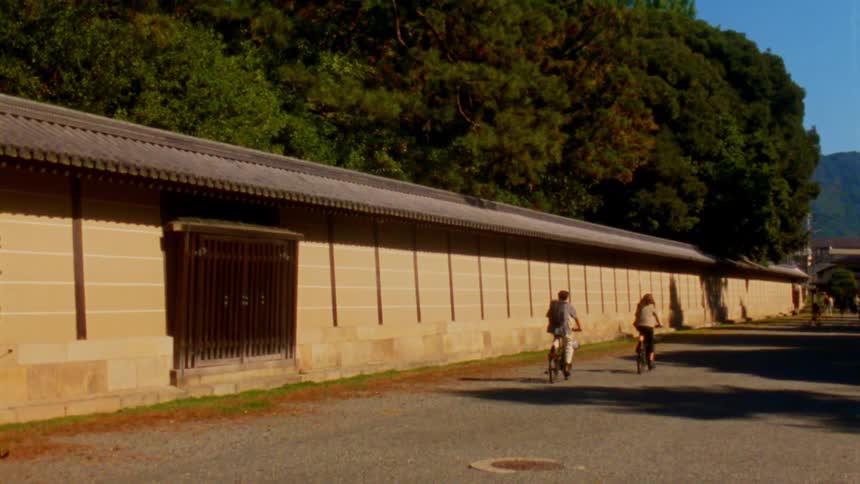 Two people are riding bicycles along a gravel path beside a long beige wall with a traditional japanese tiled roof